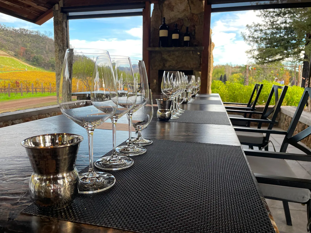 A long bar table set with multiple wine glasses and a silver ice bucket, overlooking Napa Valley vineyards and hills, with a stone fireplace in the background.
