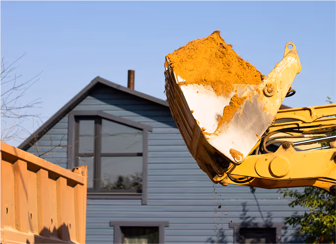 Pelleteuse jaune déchargeant des matériaux près d'une maison grise