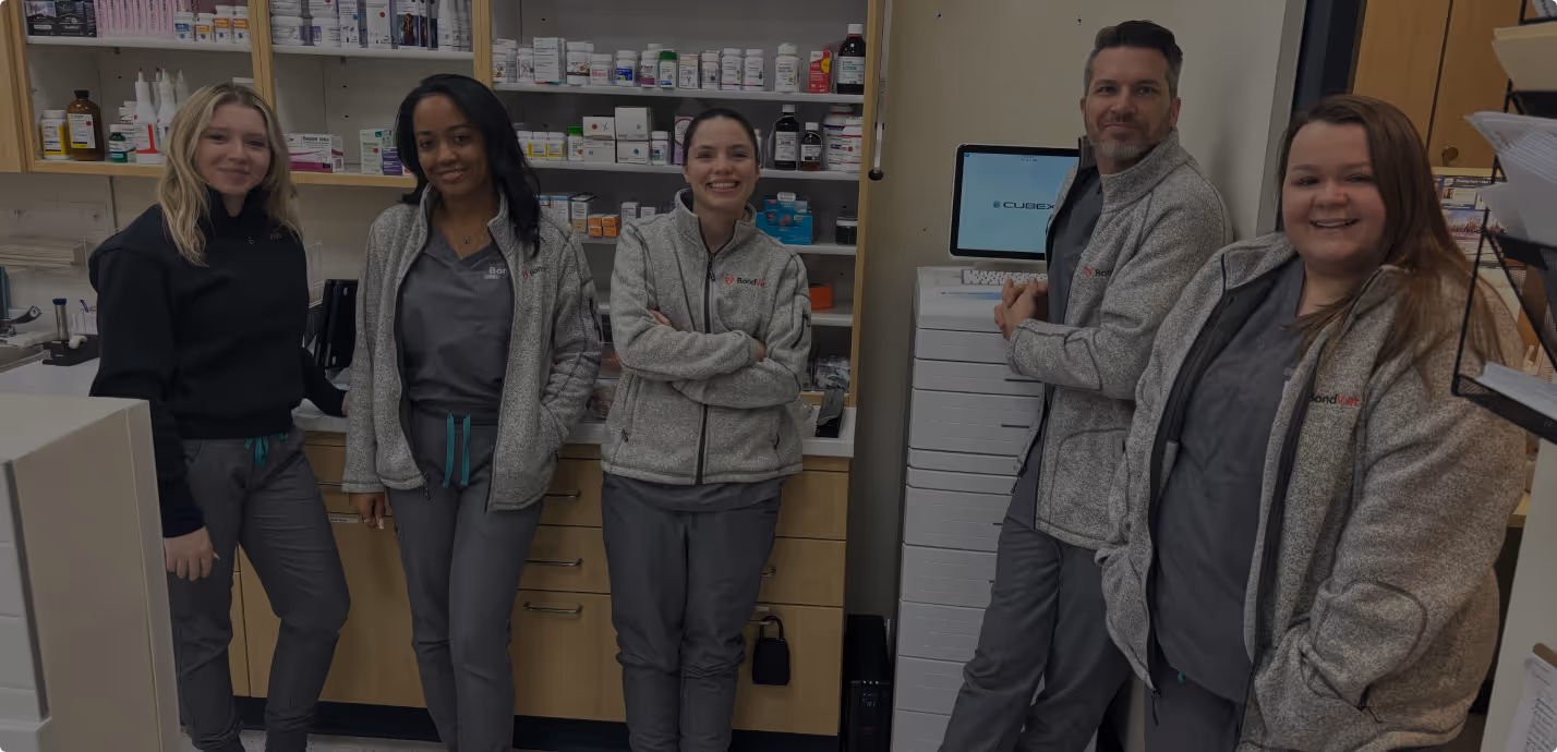 Five healthcare professionals in grey scrubs and jackets standing and smiling in a medical supply room with shelves of medicine bottles behind them.