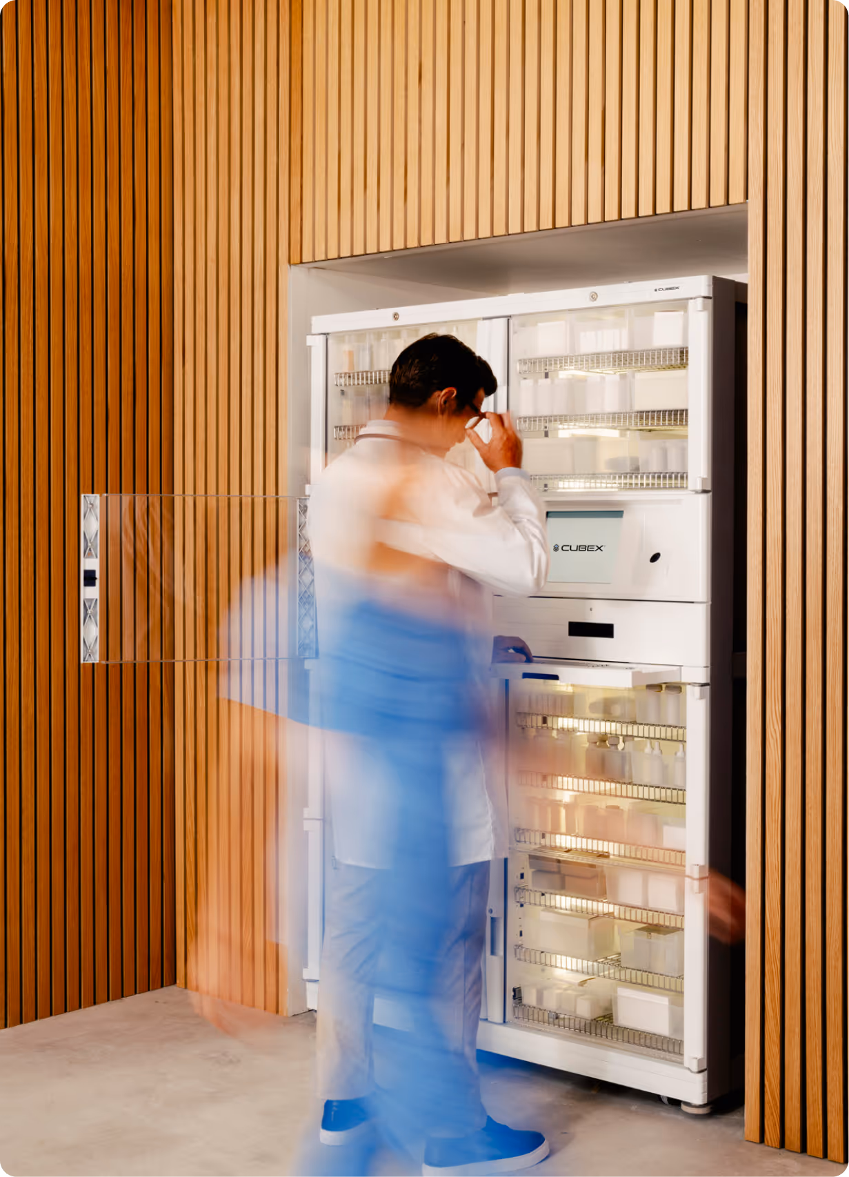Person in a white coat adjusting glasses while standing next to an open Cubex automated medicine dispenser with multiple shelves of containers, with blurred figures walking past.