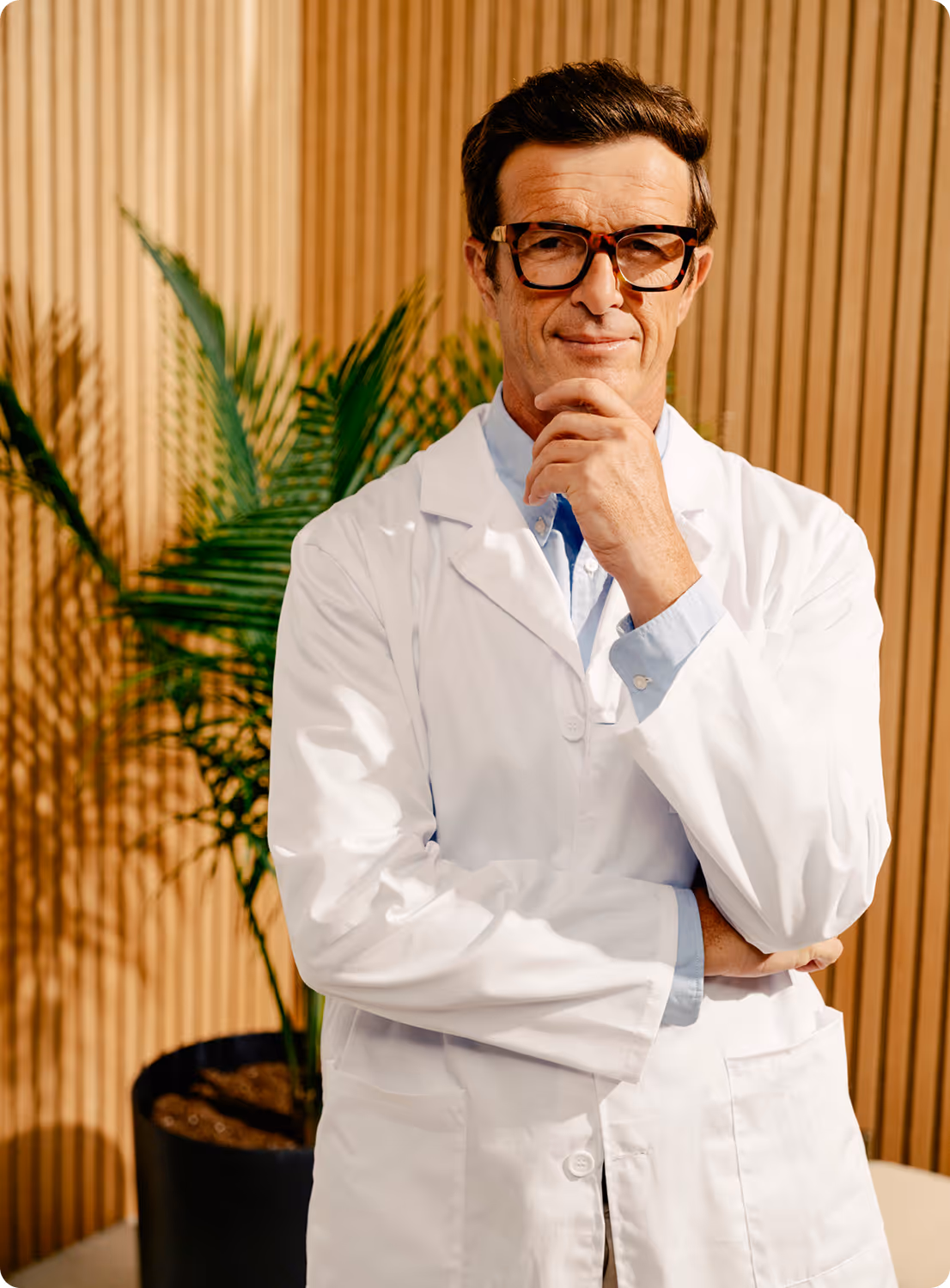 Middle-aged male doctor wearing glasses and a white lab coat, standing thoughtfully with a hand on his chin.