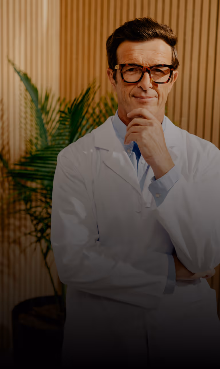 Man in glasses and white lab coat standing thoughtfully with hand on chin in front of wooden panel wall and green plant.