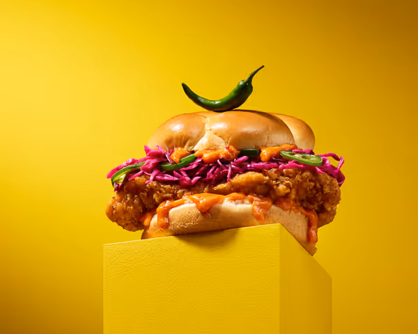 A fried chicken sandwich with red cabbage, green chili slices, and sauce sits on a yellow pedestal, topped with a whole green chili. Showcasing expert food styling and photography, it pops against the vibrant yellow background.