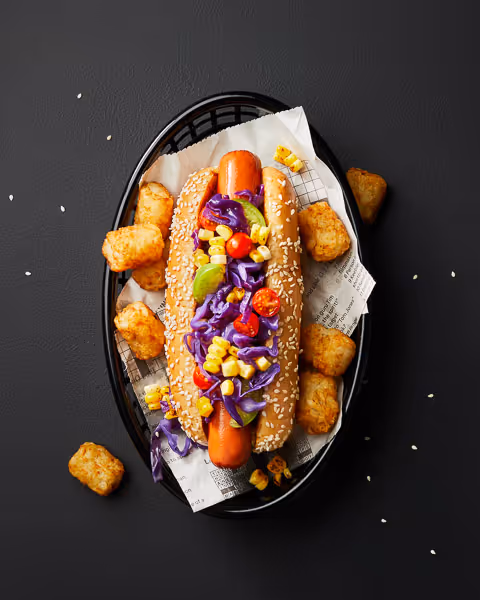 A gourmet hot dog in a sesame bun topped with purple cabbage, corn, cherry tomatoes, and avocado, served with crispy tater tots in a black basket lined with printed parchment paper on a dark textured background.