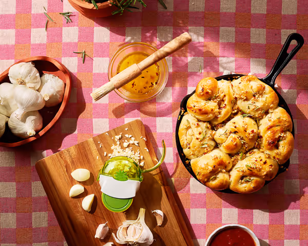 Lifestyle food photography of a skillet filled with garlic bread, surrounded by fresh garlic, herbs, and ingredients on a checkered tablecloth.