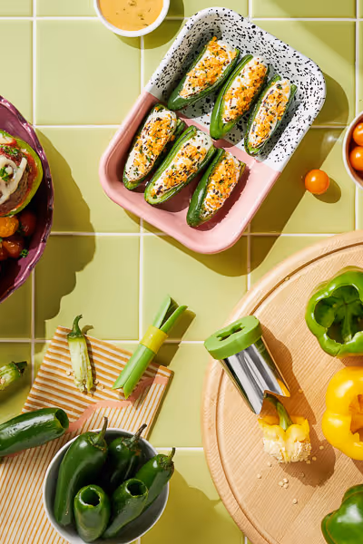 A bright kitchen scene features halved jalapeños stuffed with creamy filling in a speckled baking dish, surrounded by fresh vegetables and a jalapeño corer—perfect for the creative food lover or aspiring food photographer.