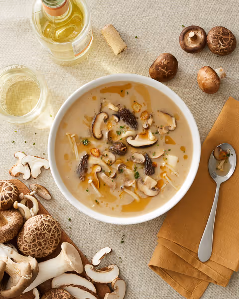 Lifestyle food photography of a hearty mushroom soup in a bowl, surrounded by fresh mushrooms, a glass of wine, and a napkin.