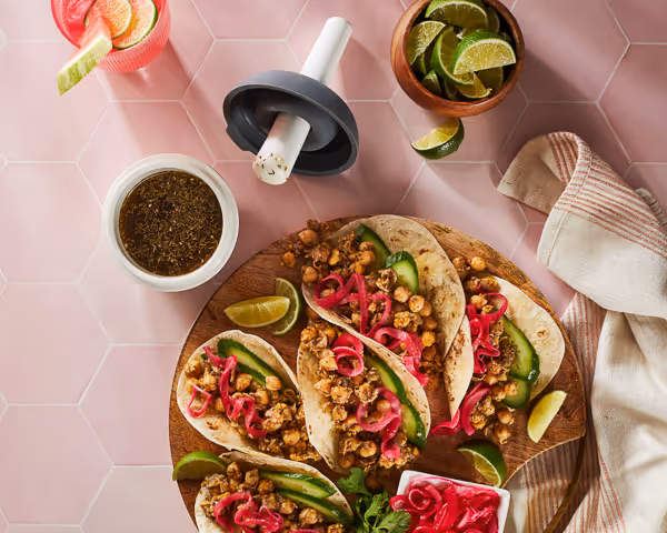 A round wooden board showcases three soft tacos with crumbled tofu or tempeh, avocado, and pink pickled onions—perfect for food photography. Lime wedges, extra pickled onions, and sauce complete the vibrant food styling on a pink hexagon-tiled surface.