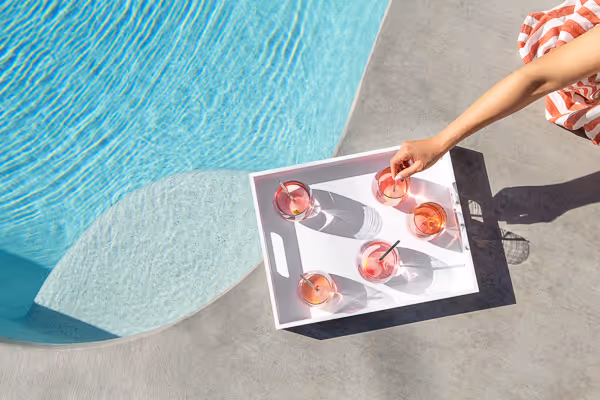 Poolside tray with six pink cocktails in sunlight and a hand reaching in.