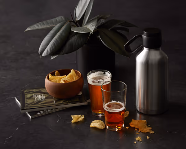Two glasses of amber beer with chips and a stainless steel growler on a black marble table.