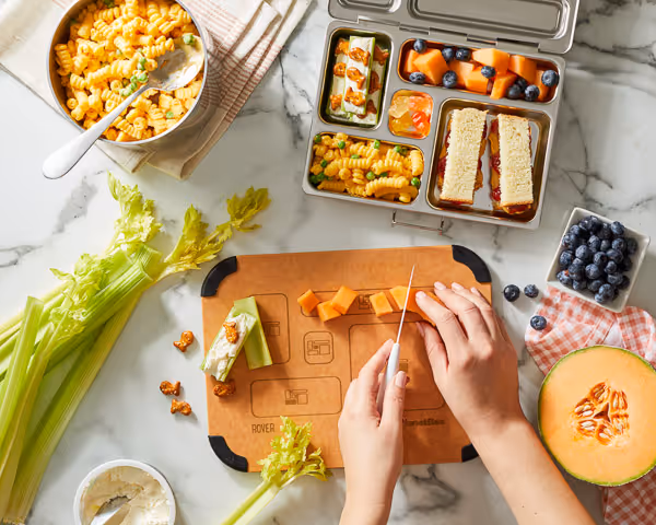 Overhead view of a child’s lunchbox being prepared with cantaloupe, blueberries, celery, and pasta on a marble countertop