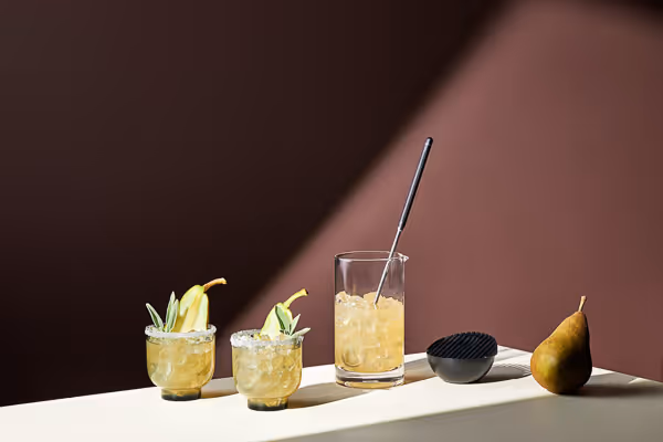 Beverage studio photography with two garnished cocktails, a mixing glass, and a pear on a light surface against a dark background.