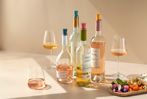 Hard light food photography of wine bottles and glasses with different wines on a light surface, accompanied by a plate of fresh vegetables.