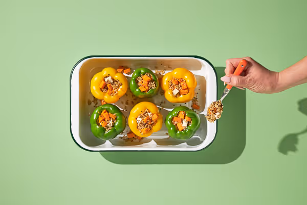 Top view of stuffed bell peppers in a baking dish, with a hand adding grains using a spoon.
