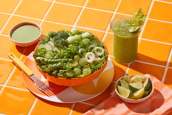 Studio food photography of a monochromatic green salad in an orange bowl, with a green smoothie and dressing on orange tiles.