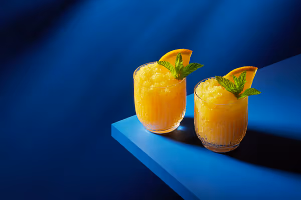 Cocktail photography of two vibrant orange vodka slush drinks garnished with orange slices and mint, set against a deep blue background.
