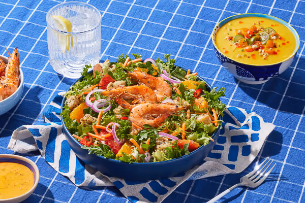 Overhead lifestyle food shot of a vibrant shrimp salad with fresh greens and tomatoes in a blue bowl, gazpacho, and lemon water.
