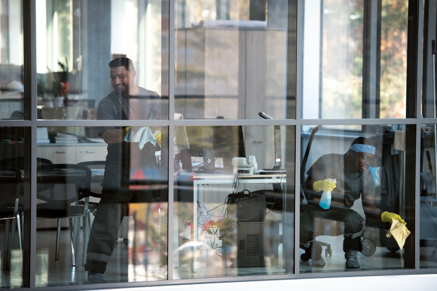 Two janitors cleaning desks and windows inside an office with large glass panels.