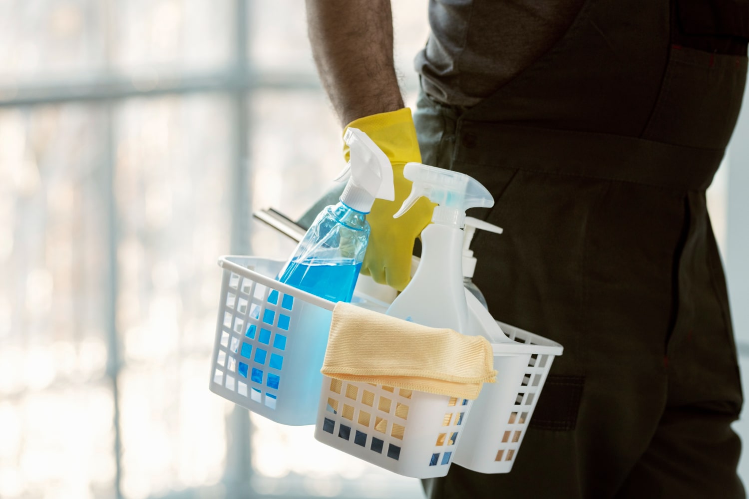 Person wearing yellow gloves holding a white basket with cleaning spray bottles and a yellow cloth.