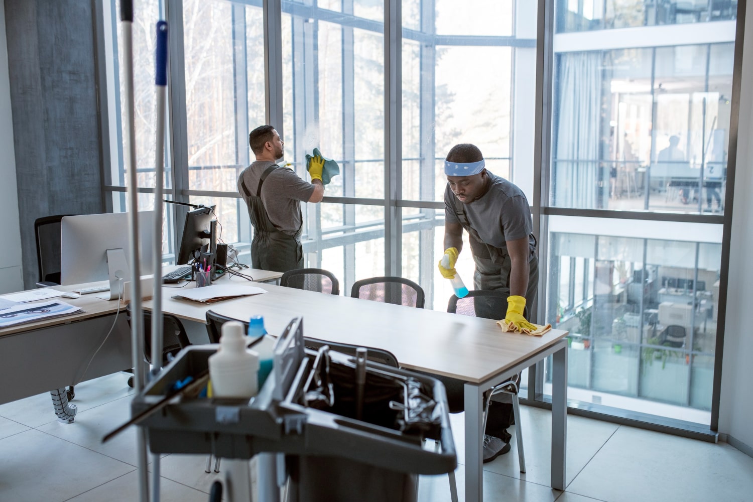 Two janitors cleaning a modern office with large windows, one wiping a table and the other cleaning glass panels.