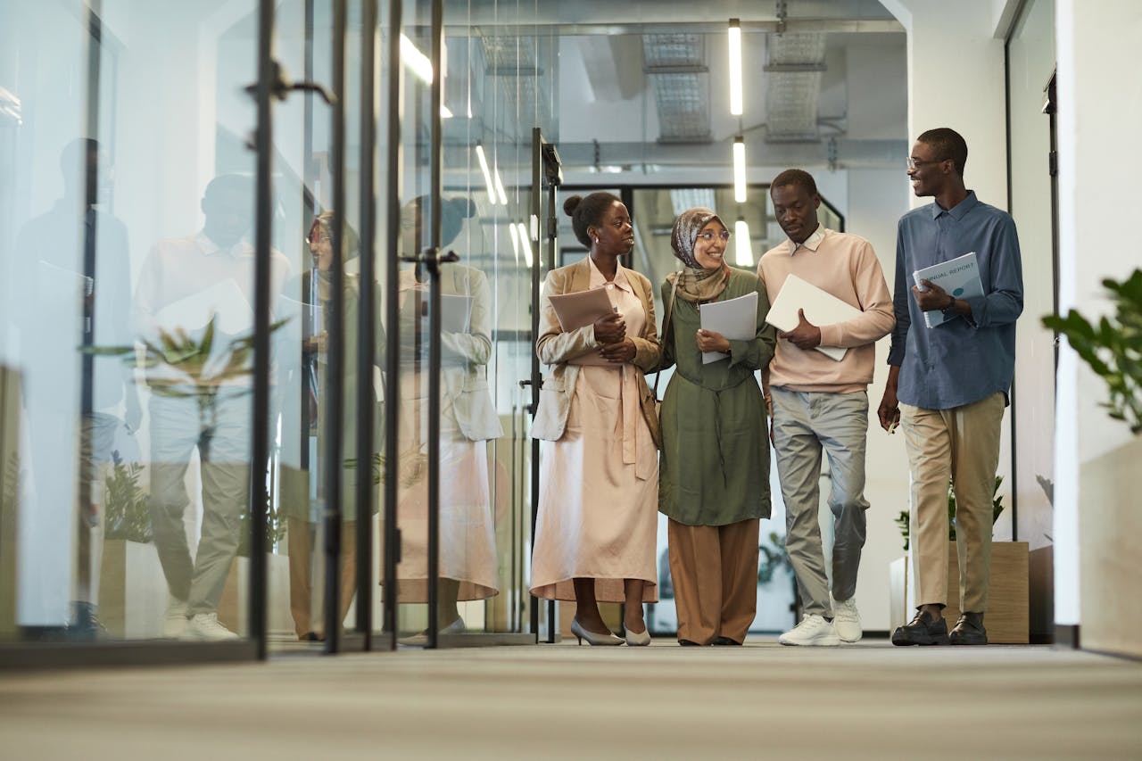 Four diverse colleagues walking and talking in a modern office hallway holding laptops and documents.