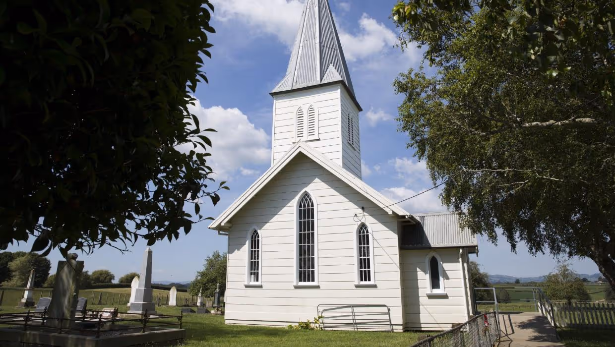 A white wooden church with a tall steeple, surrounded by greenery and gravestones, under a blue sky with fluffy clouds.