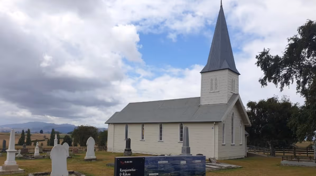 A quaint white wooden church with a tall spire, surrounded by gravestones and green lawns under a cloudy sky.