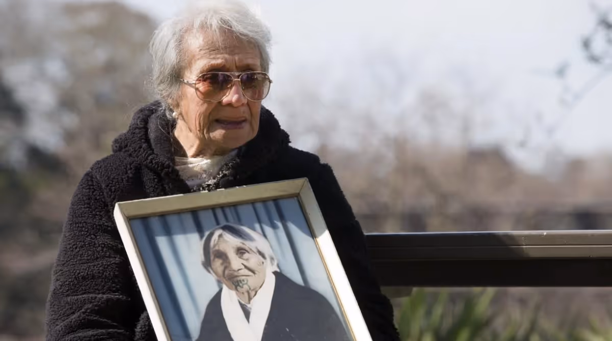 Elderly woman in sunglasses holds a framed photo of another woman, outdoors with trees blurred in the background.