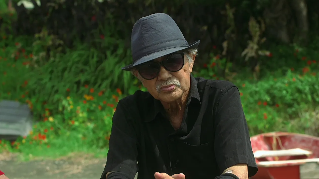 An older man sits outdoors, wearing a black shirt, sunglasses, and a fedora, with greenery and flowers in the background.