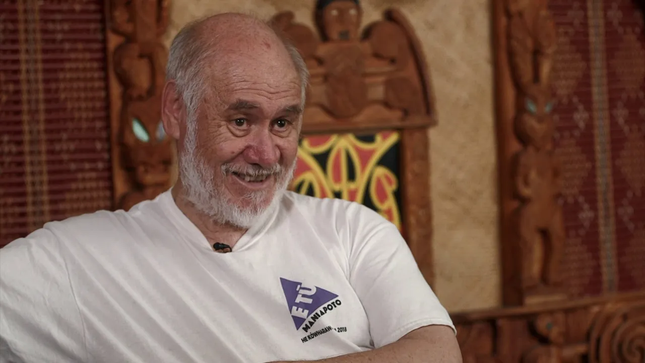 A smiling man with a white beard sits against a backdrop of traditional carvings, wearing a plain white shirt.