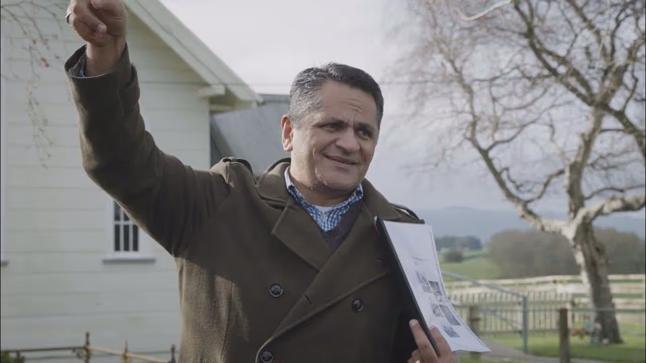 A man in a coat is gesturing with one hand while holding a document. A pastoral landscape and a house are in the background.