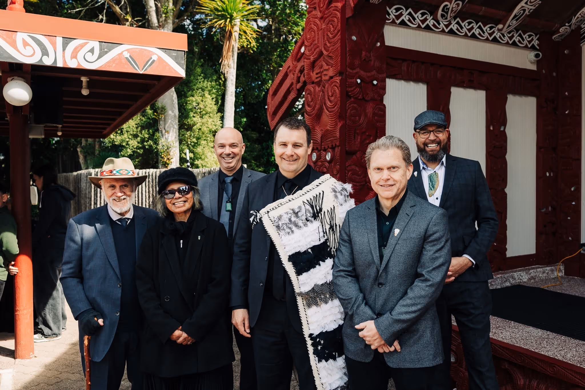 Group of six people in formal attire posing outdoors near a traditional patterned structure, with trees in the background.