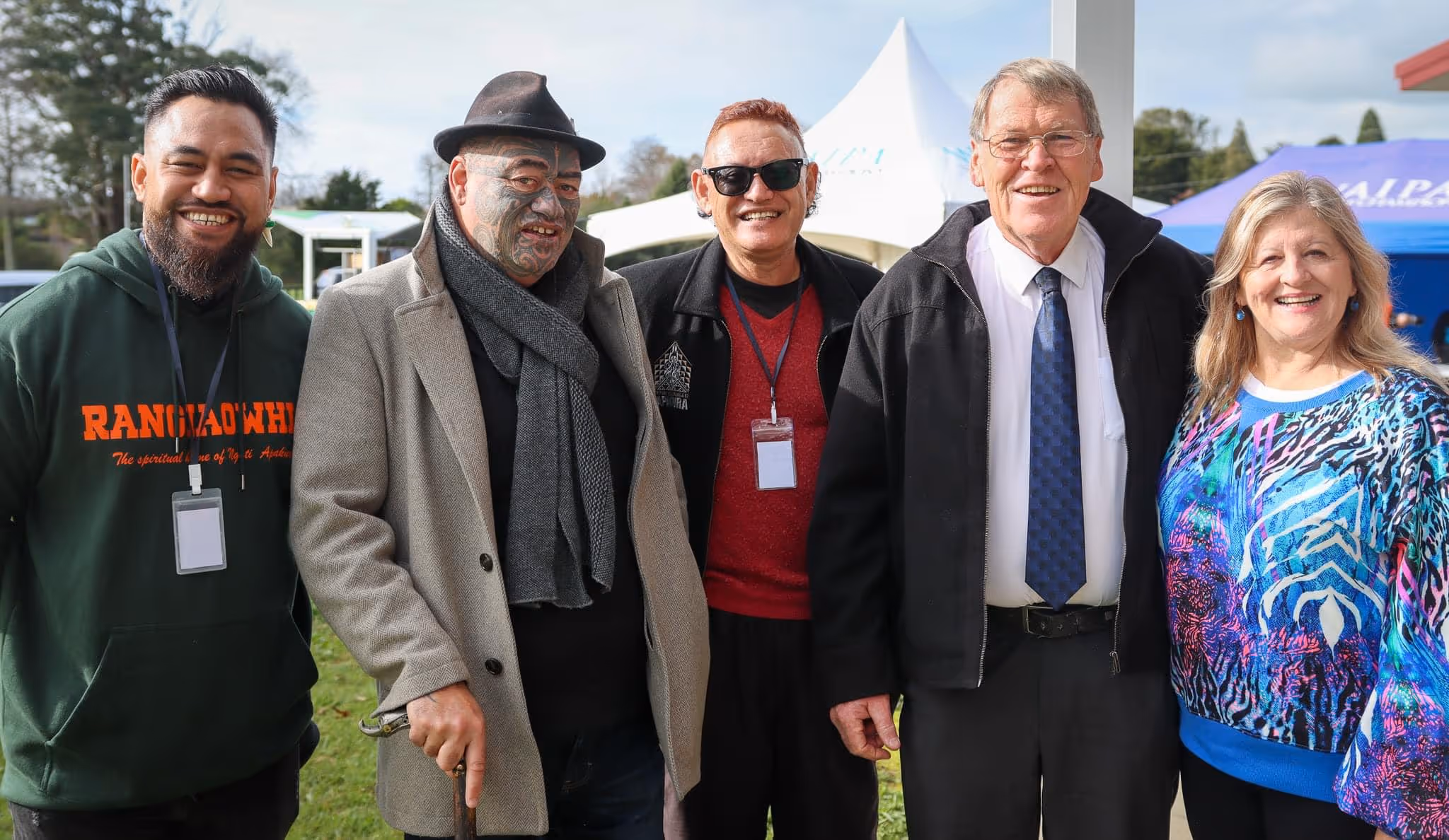 Five individuals stand together outdoors, smiling, dressed in casual and formal attire, with a canopy and trees in the background.