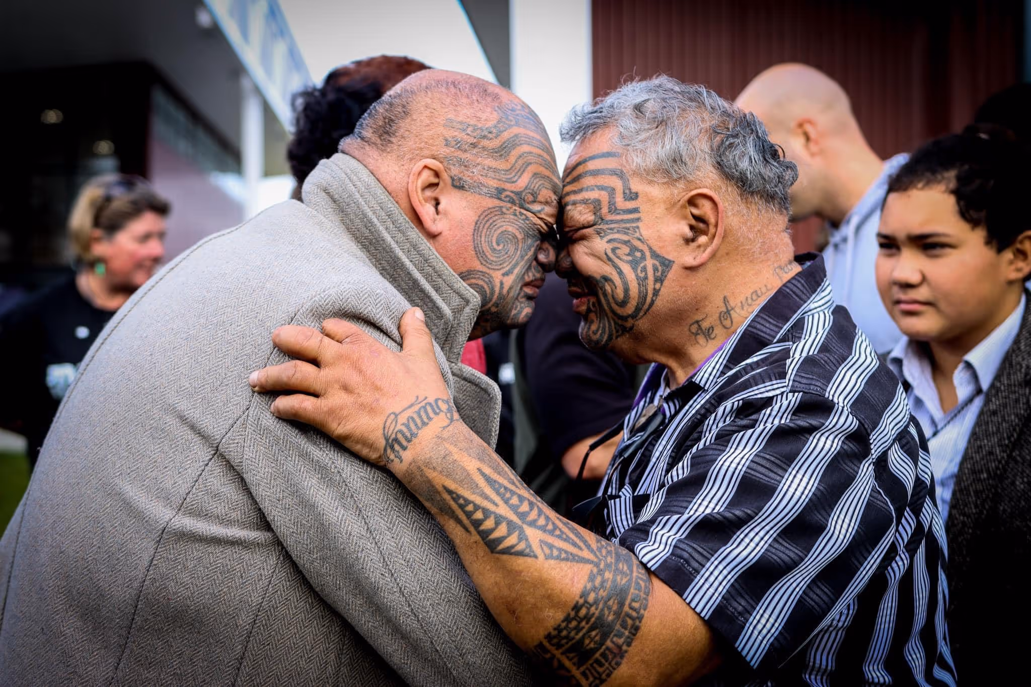 Two men with traditional tattoos share a hongi (forehead greeting) while embracing, surrounded by others in a culturally meaningful setting.