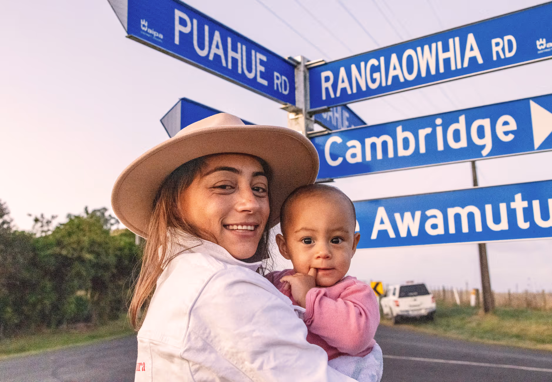 A woman wearing a hat smiles while holding a baby at a roadside signpost with directional signs for Puahue, Rangiaowhia, Cambridge, and Awamut.