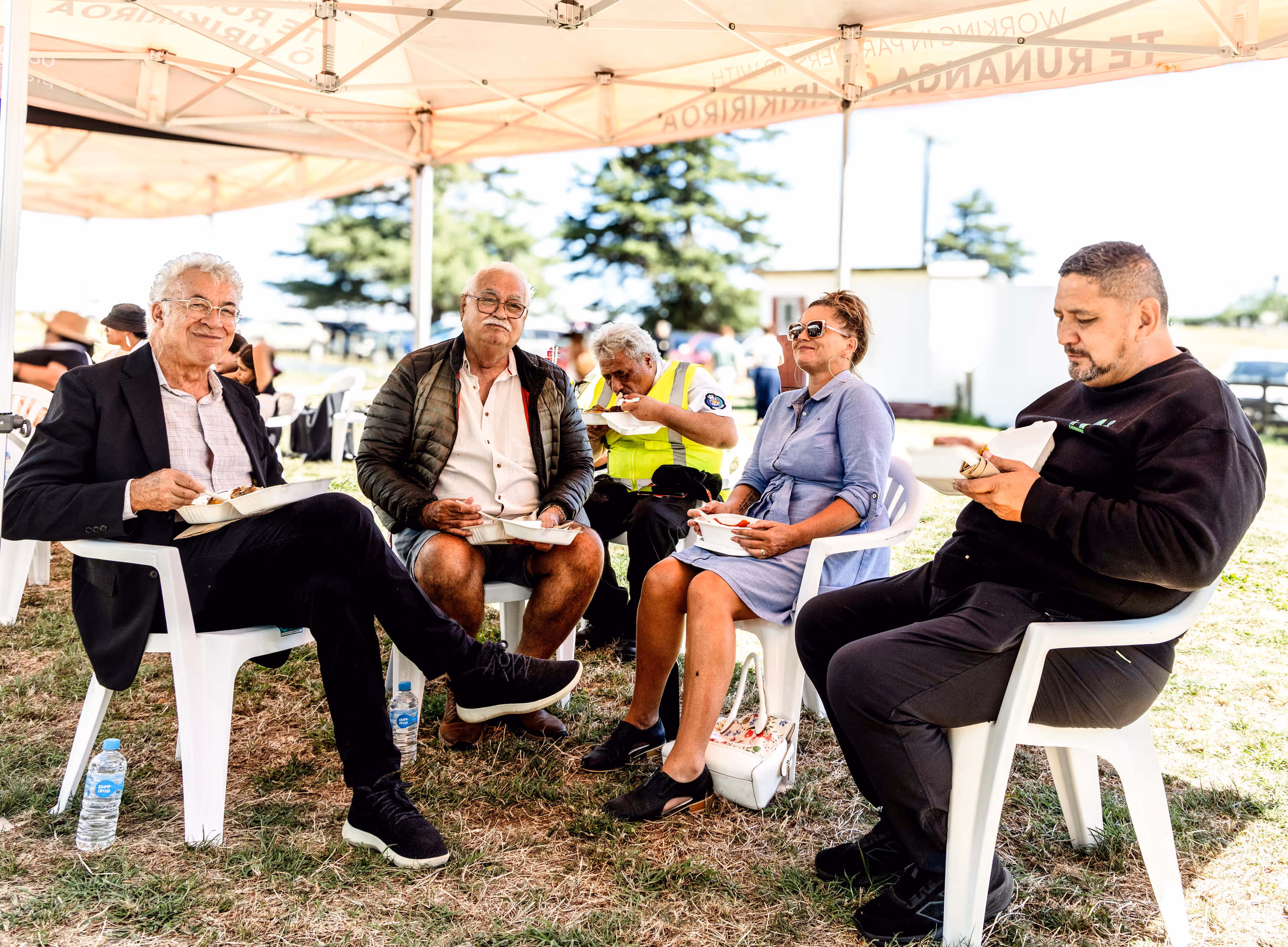 Five people sit under a canopy, enjoying food. Two men wear casual attire, a woman in a dress, and two others relax nearby. Green grass surrounds them.