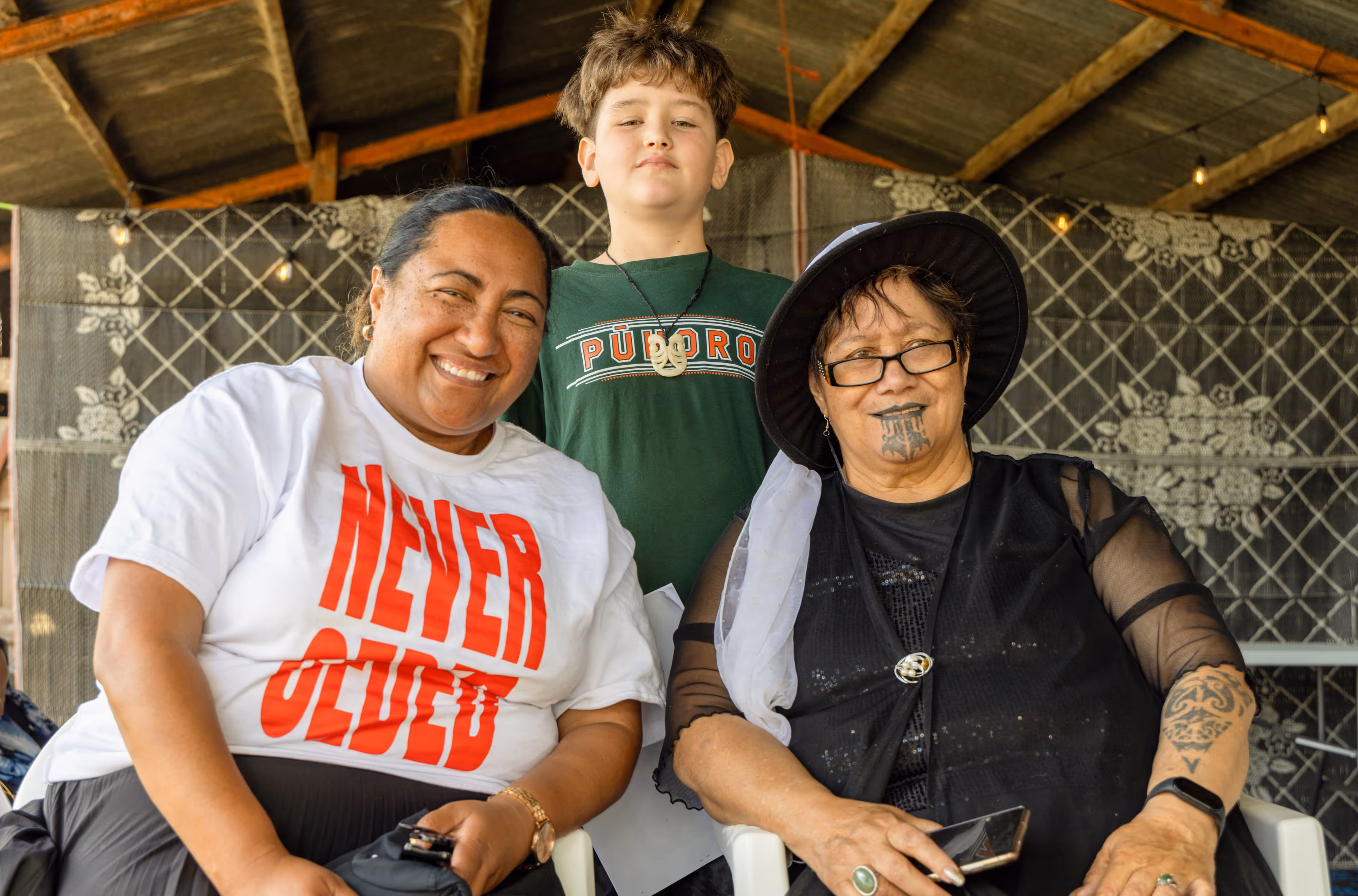 Three individuals smile together while sitting in a shaded area. The woman on the left wears a white shirt with red text, and the woman on the right has a black hat.