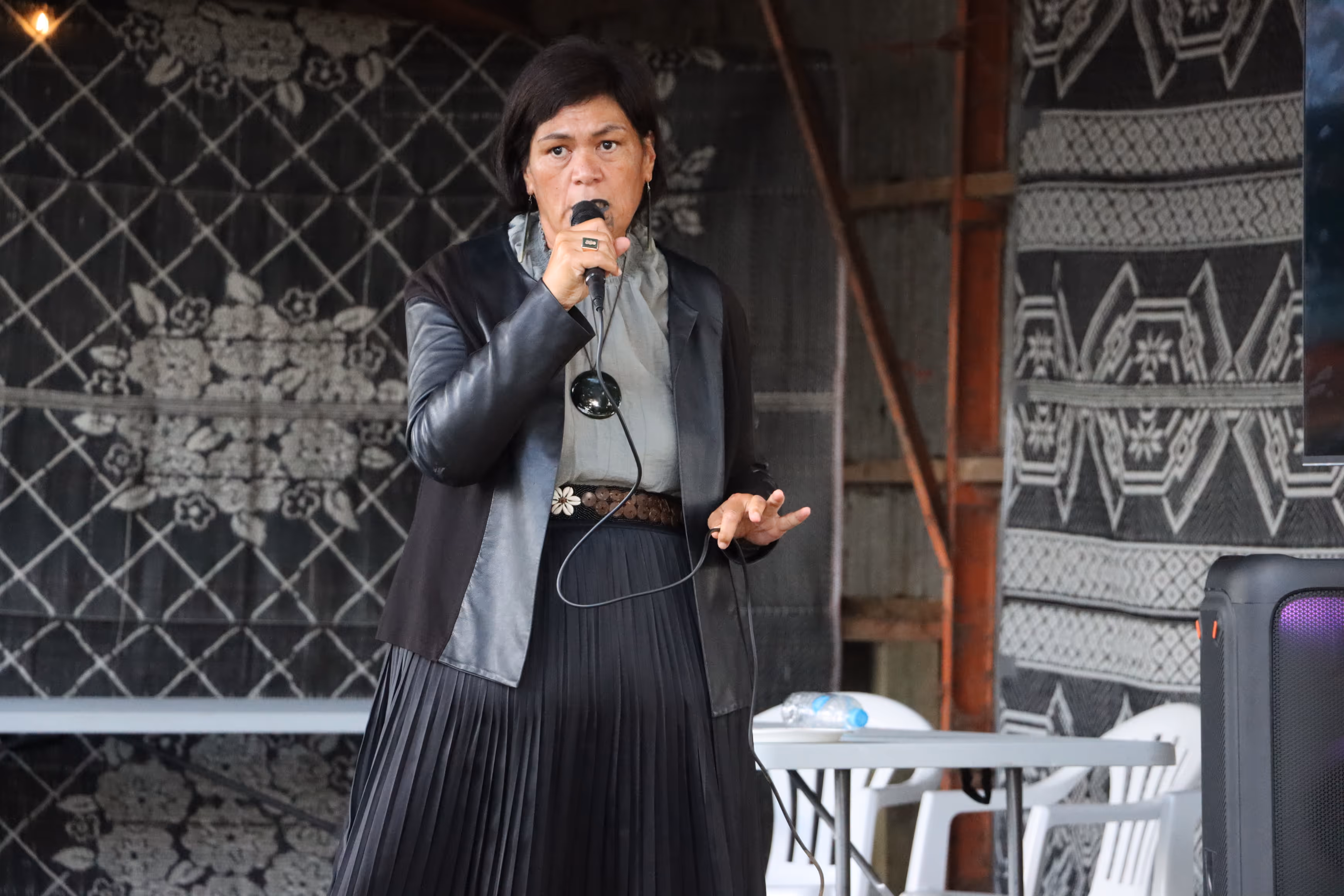 A woman with shoulder-length dark hair speaks into a microphone, wearing a black leather jacket and a pleated skirt, against a patterned background.