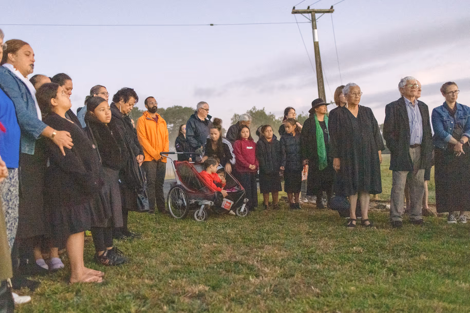 A diverse group of people, including adults and children, gather solemnly outdoors at dusk, some wearing black attire, with a child in a stroller.