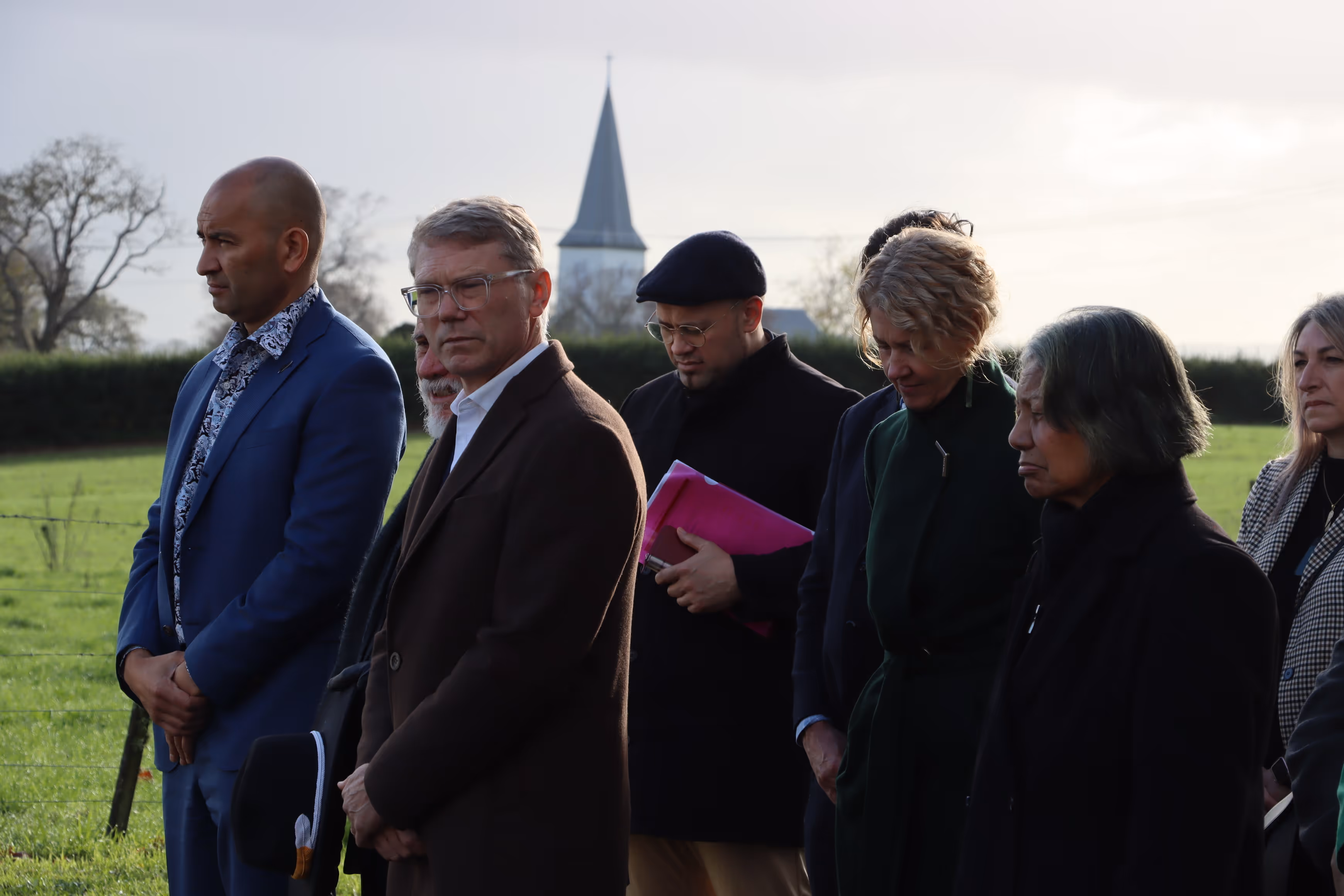 A group of people stands in a field, some with heads bowed, while a church spire is visible in the background.