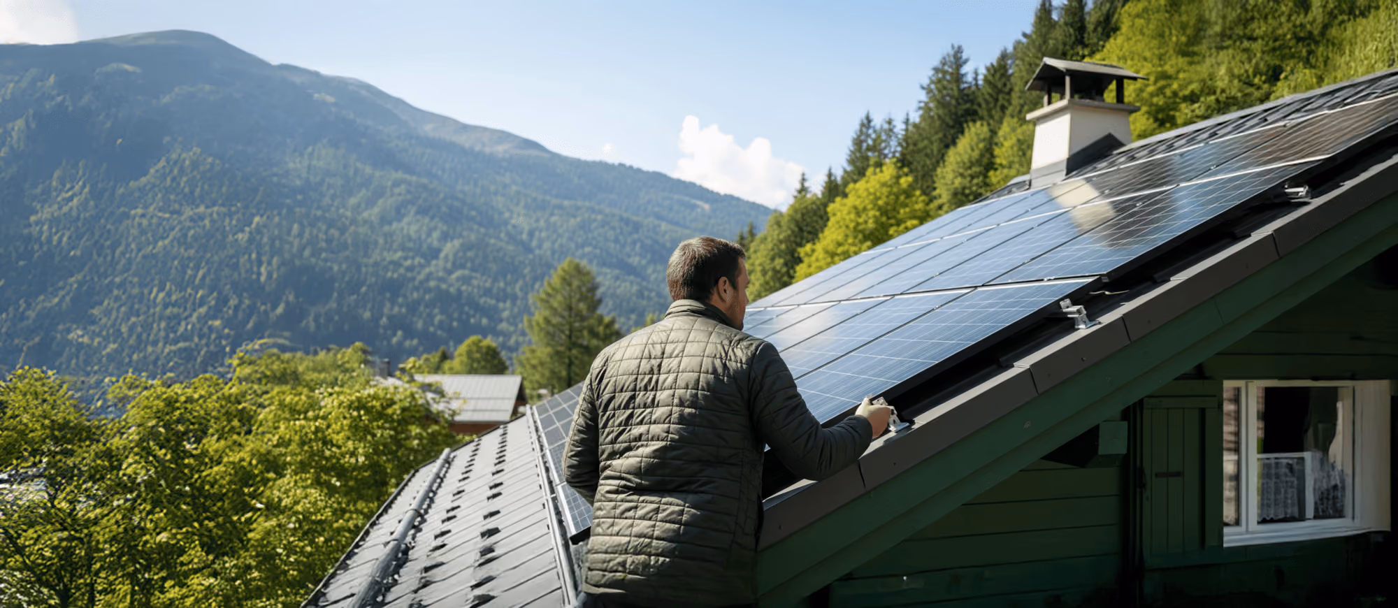 Un homme en veste matelassée inspecte des panneaux solaires sur le toit d'une maison en bois entourée de forêts et de montagnes.