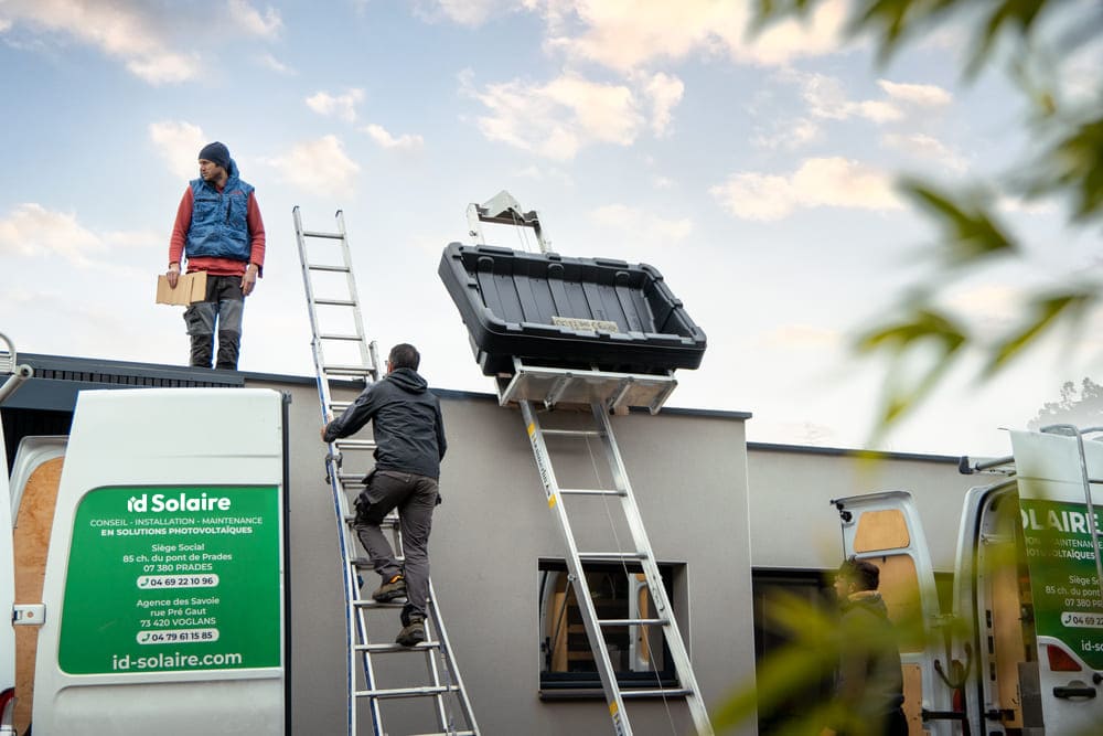Deux ouvriers utilisent des échelles pour accéder au toit d'un bâtiment, avec un monte-charge installé pour transporter des matériaux, à côté de deux véhicules de service ID Solaire.