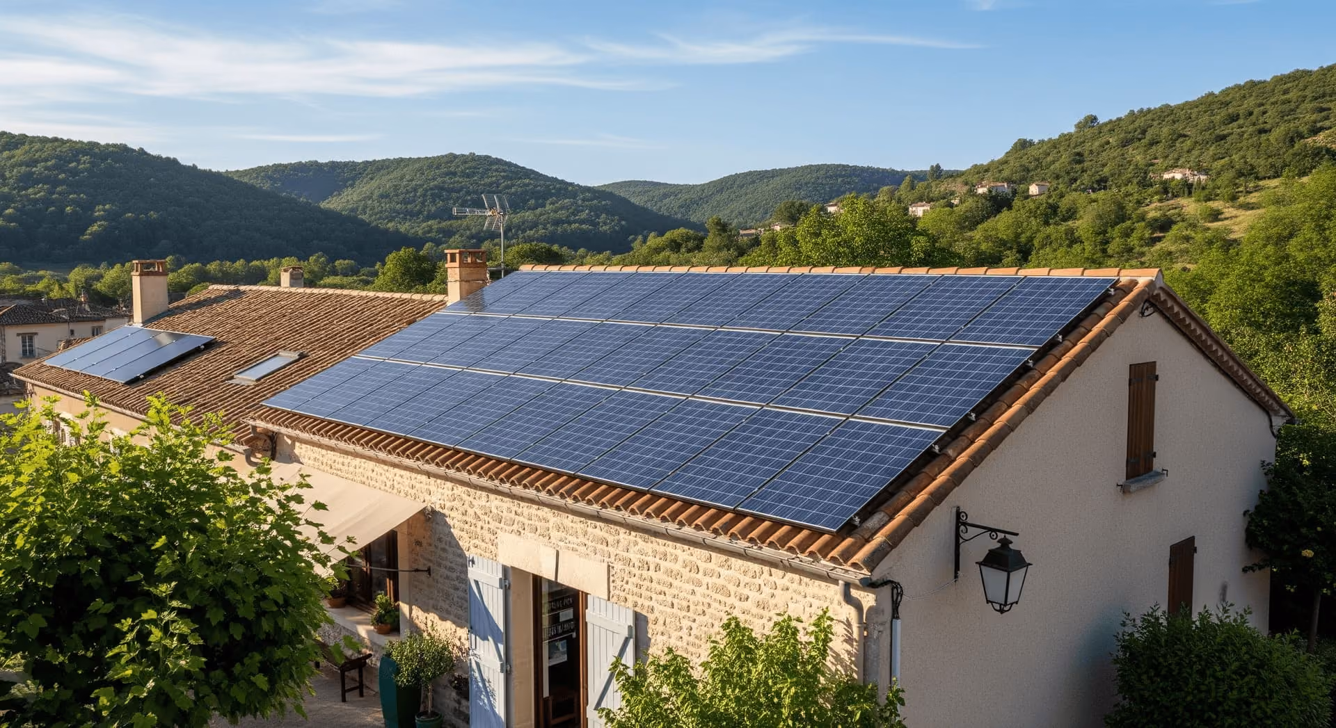 Maison en pierre avec plusieurs panneaux solaires installés sur un toit en tuiles, entourée de verdure et de collines au coucher du soleil.