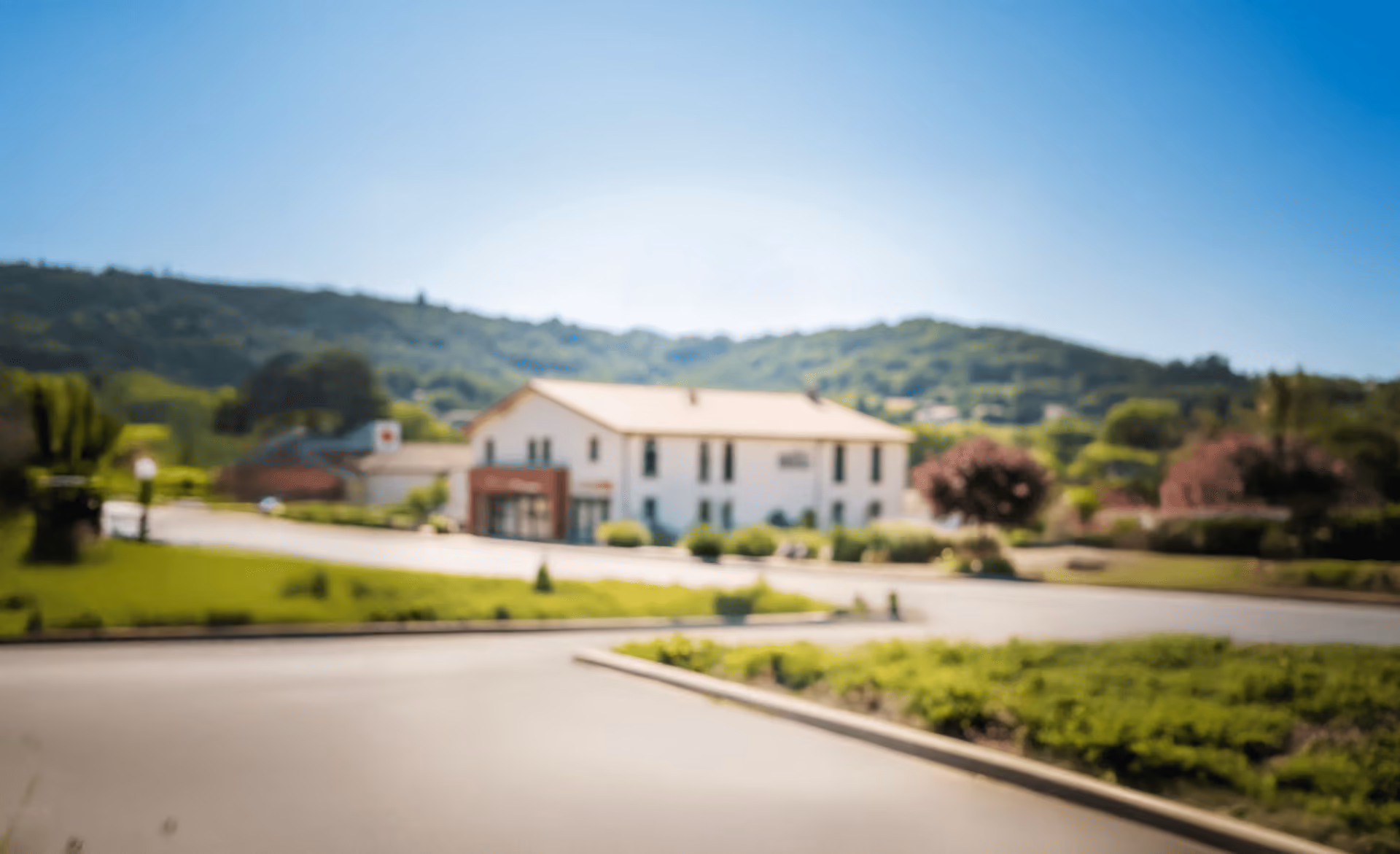 Bâtiment blanc flou avec un toit beige entouré de verdure et de collines sous un ciel bleu clair.