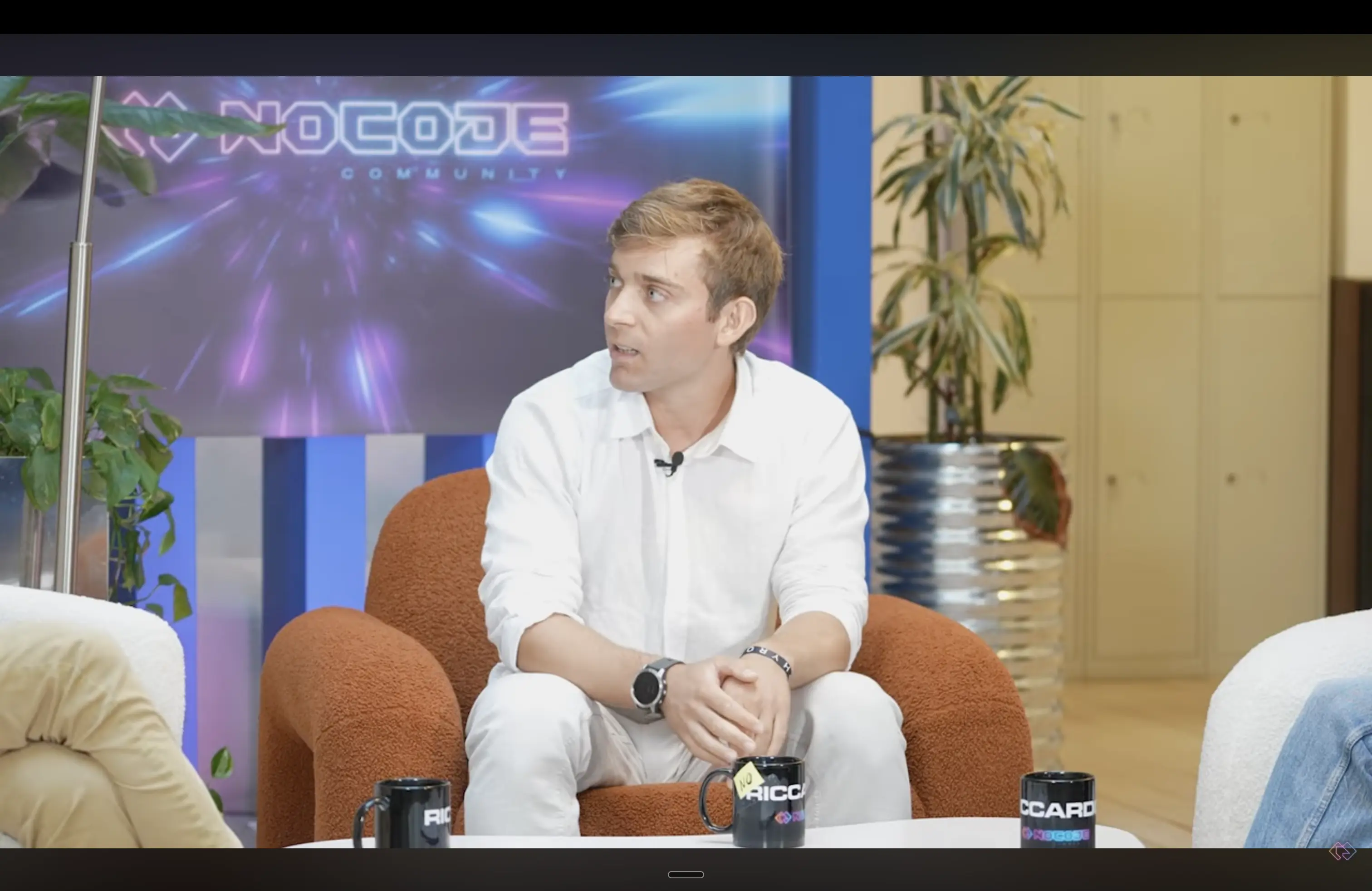 Man in a white shirt sitting on a brown chair during an indoor interview with 'NocojE Community' neon sign in the background.