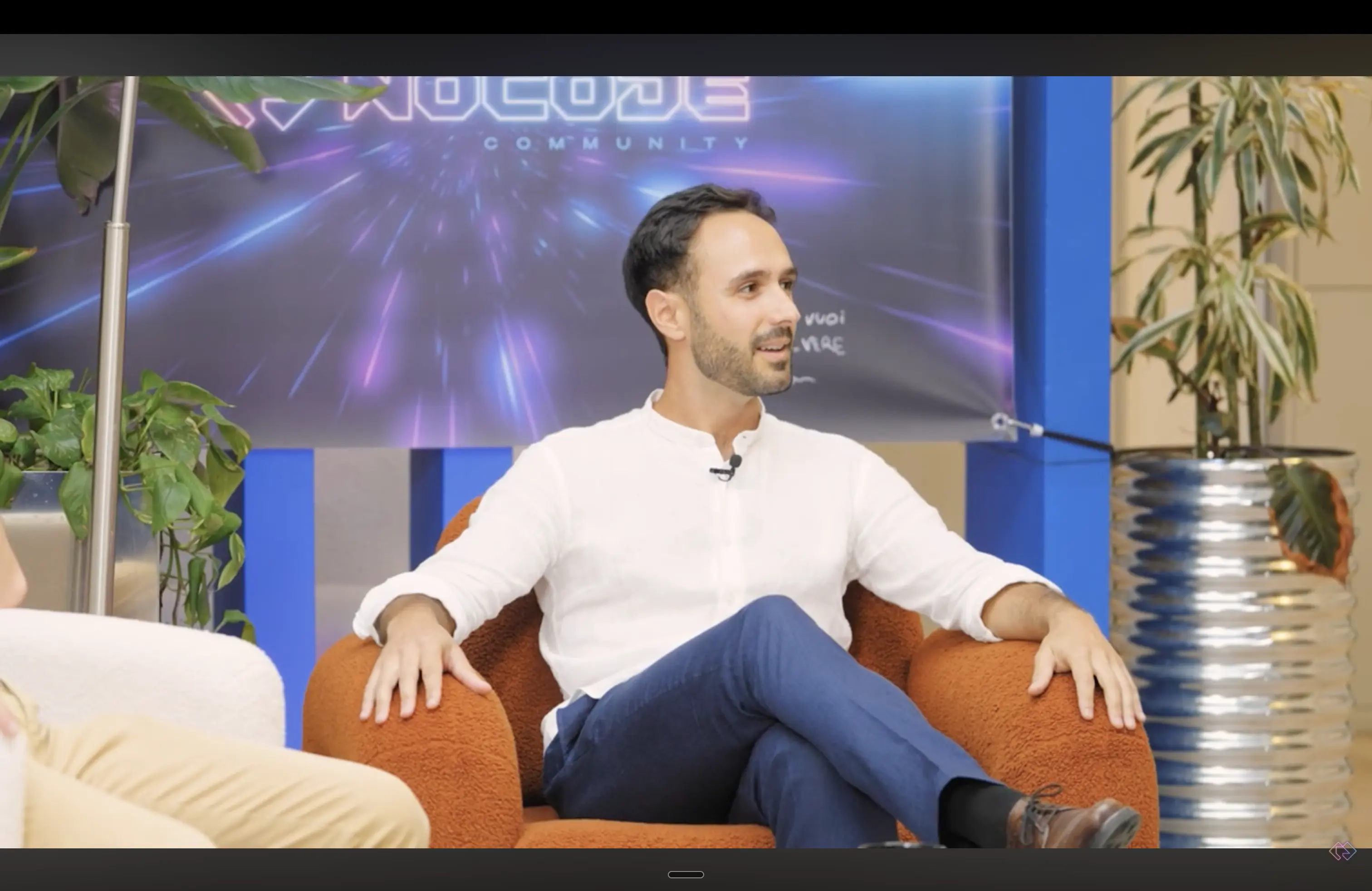 Man in a white shirt and blue pants sitting cross-legged on an orange chair with plants and a neon-lit backdrop behind him.