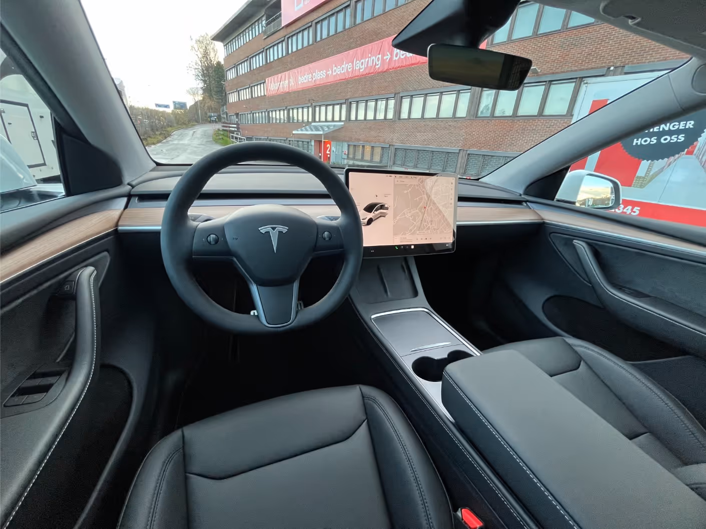 Interior view of a Tesla car showing a black steering wheel with Tesla logo, a central touchscreen display, and black leather seats with wooden trim on the dashboard.