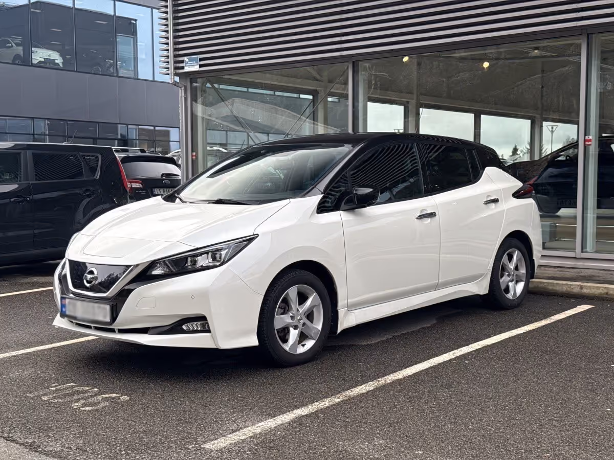 White Nissan Leaf electric car parked in a lot beside a modern building with large windows.