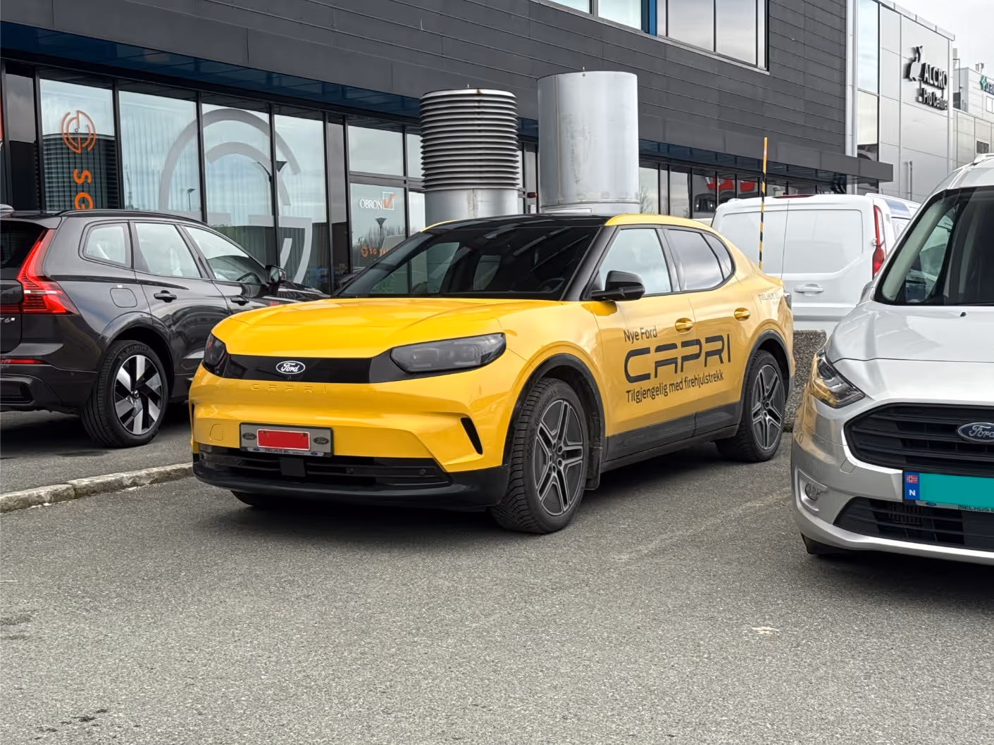 Yellow Ford Capri car with black accents parked between a black SUV and a white van in front of a commercial building.