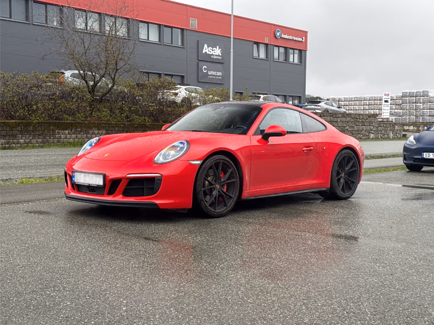Red Porsche sports car parked on wet asphalt in front of a gray and red building with company signs.